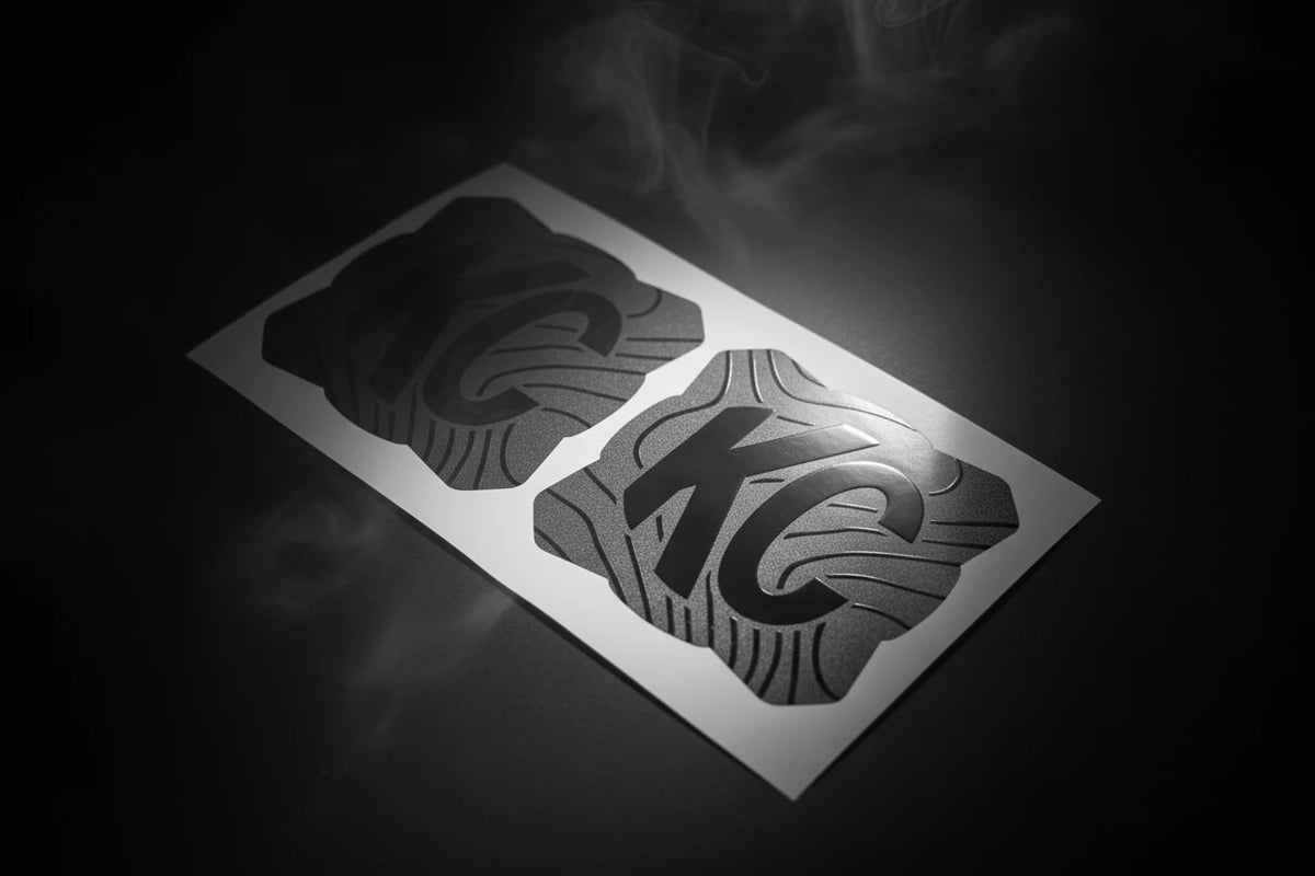 Two black and gray 'KC' stickers on a white card with dark background